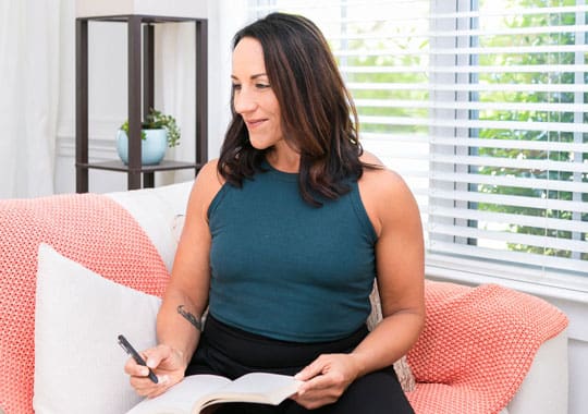 Jess Beran, Charleston SC intuitive life coach, sitting on a couch with a journal and phone in hand, reflecting in a bright and welcoming coaching space