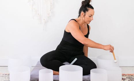 Jess Beran leading a sound bowl meditation session, seated on the floor and gently playing crystal singing bowls in a calm, minimalist space