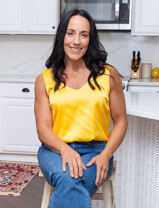 Jess Beran, Charleston SC intuitive life coach, smiling in a bright kitchen setting wearing a yellow sleeveless blouse and jeans, professional headshot for her coaching brand
