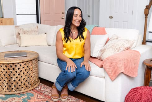 Jess Beran with long dark hair wearing a bright yellow sleeveless top and jeans, sitting on a light-colored couch in a cozy living room with a wooden coffee table and laptop nearby